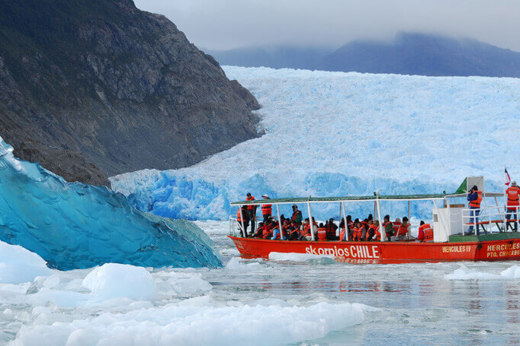 A small red excursion boat from an Australis Cruise navigates through icy waters near a massive blue glacier wall. 