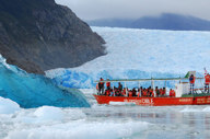 A small red excursion boat from an Australis Cruise navigates through icy waters near a massive blue glacier wall. 