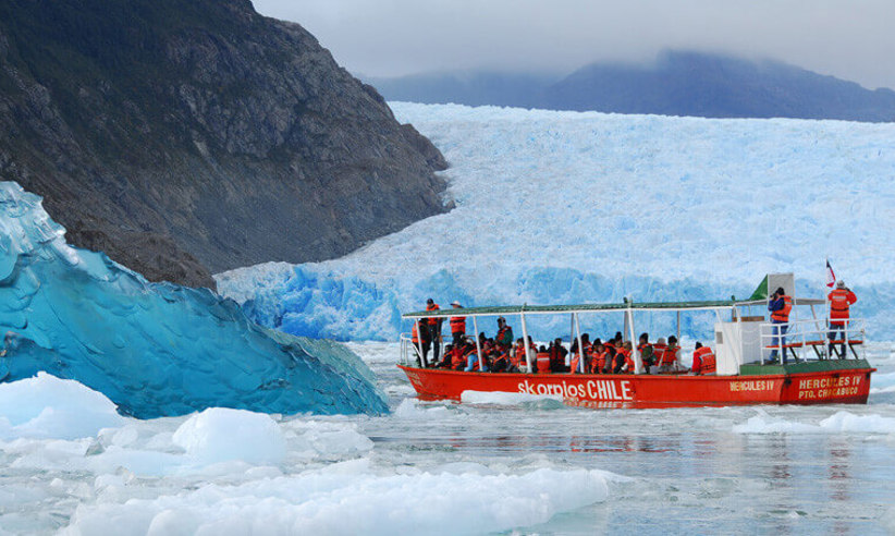 A small red excursion boat from an Australis Cruise navigates through icy waters near a massive blue glacier wall. 
