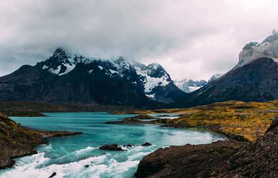 Turquoise glacial waters reflect rugged, snow-capped peaks beneath a moody sky in Torres del Paine National Park, Chile. 