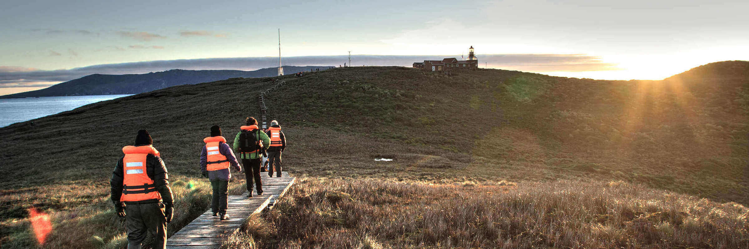 Travellers walk toward the Cape Horn lighthouse at sunrise, surrounded by rugged Patagonian terrain and ocean views. 