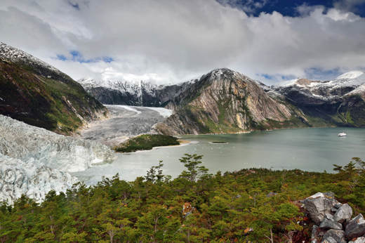 Dramatic view of a glacier flowing into a coastal fjord, framed by lush forest and rugged mountains in southern Chile. 