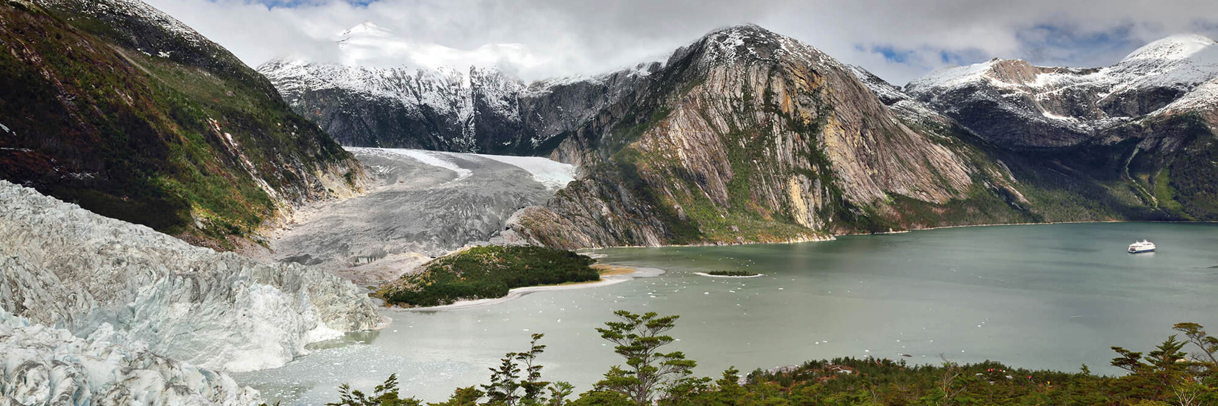 Dramatic view of a glacier flowing into a coastal fjord, framed by lush forest and rugged mountains in southern Chile. 