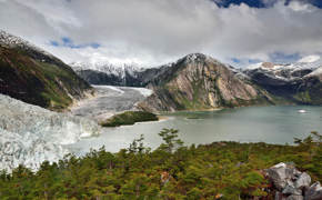Dramatic view of a glacier flowing into a coastal fjord, framed by lush forest and rugged mountains in southern Chile. 