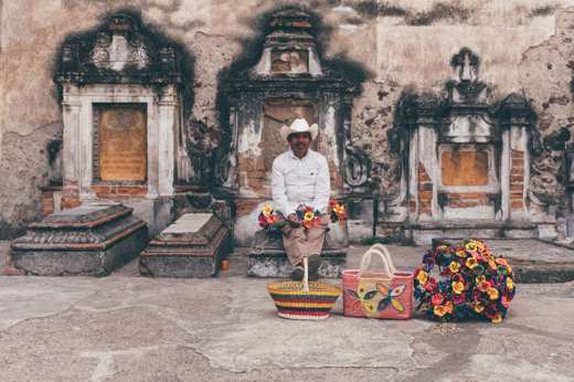 Mexican street vendor with colourful flowers and baskets sits by a rustic colonial wall, capturing local charm and heritage. 