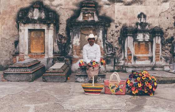 Mexican street vendor with colourful flowers and baskets sits by a rustic colonial wall, capturing local charm and heritage. 