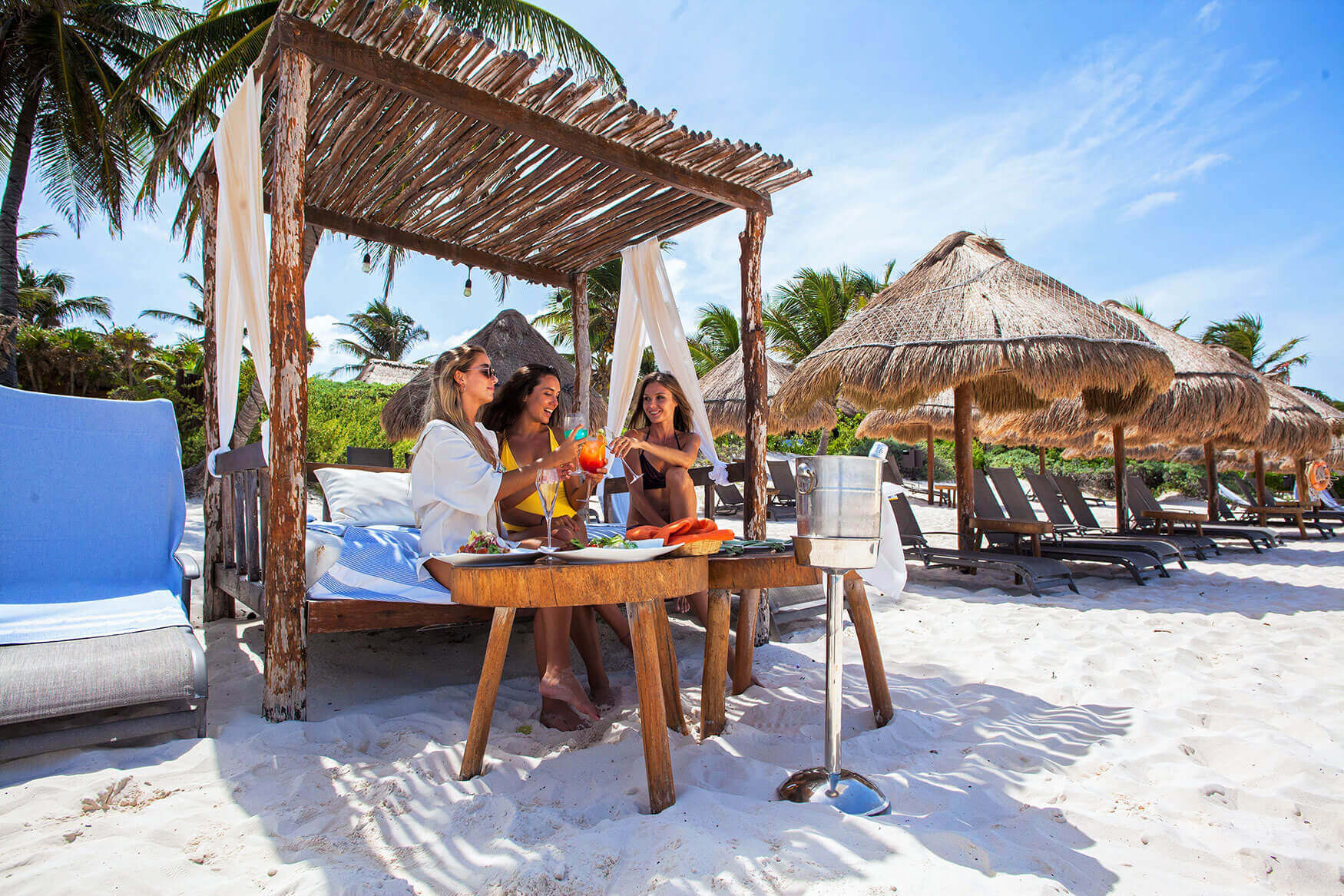 Guests enjoying drinks under a shaded beach cabana at a luxury hotel in Tulum, Mexico, with palm trees and white sand. 