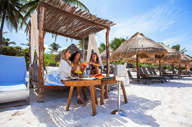 Guests enjoying drinks under a shaded beach cabana at a luxury hotel in Tulum, Mexico, with palm trees and white sand. 