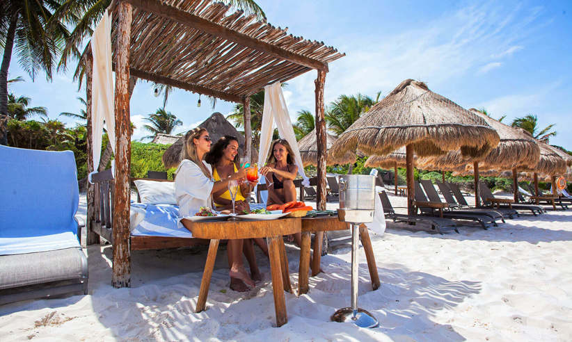 Guests enjoying drinks under a shaded beach cabana at a luxury hotel in Tulum, Mexico, with palm trees and white sand. 