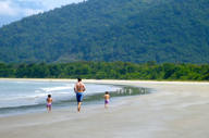 Father and children walking along the tranquil beach of Angra dos Reis, Brazil with lush forested hills in the background. 