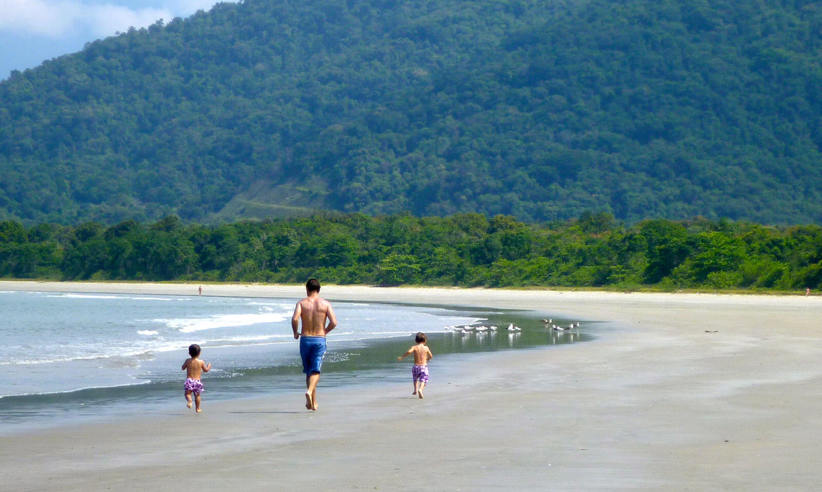 Father and children walking along the tranquil beach of Angra dos Reis, Brazil with lush forested hills in the background. 