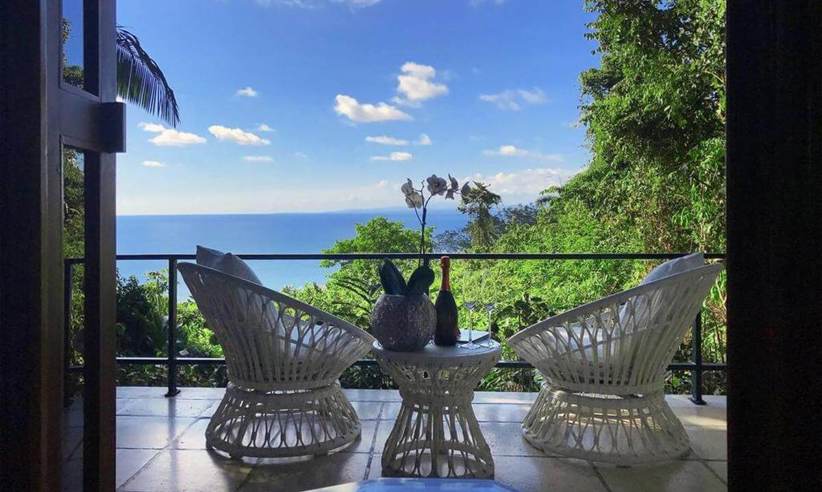 Private balcony with elegant white wicker chairs overlooking lush greenery and the Pacific Ocean. 