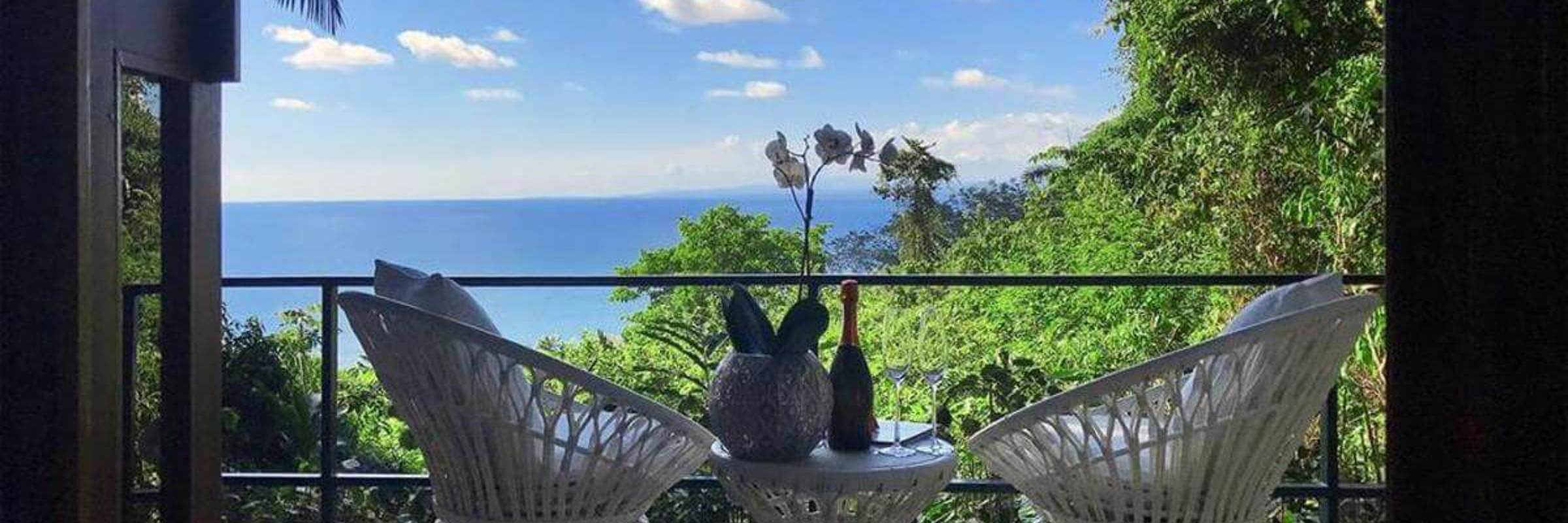 Private balcony with elegant white wicker chairs overlooking lush greenery and the Pacific Ocean. 