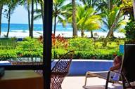 A tourist lounges on a terrace with a view of palm trees, a pool, and Playa Esterillos Beach in Costa Rica. 