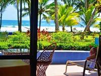 A tourist lounges on a terrace with a view of palm trees, a pool, and Playa Esterillos Beach in Costa Rica. 