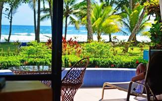 A tourist lounges on a terrace with a view of palm trees, a pool, and Playa Esterillos Beach in Costa Rica. 