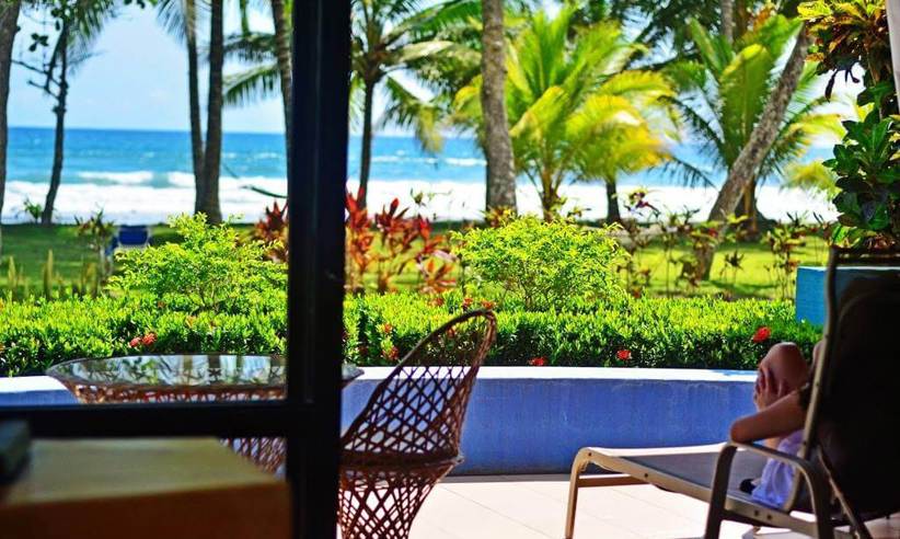 A tourist lounges on a terrace with a view of palm trees, a pool, and Playa Esterillos Beach in Costa Rica. 