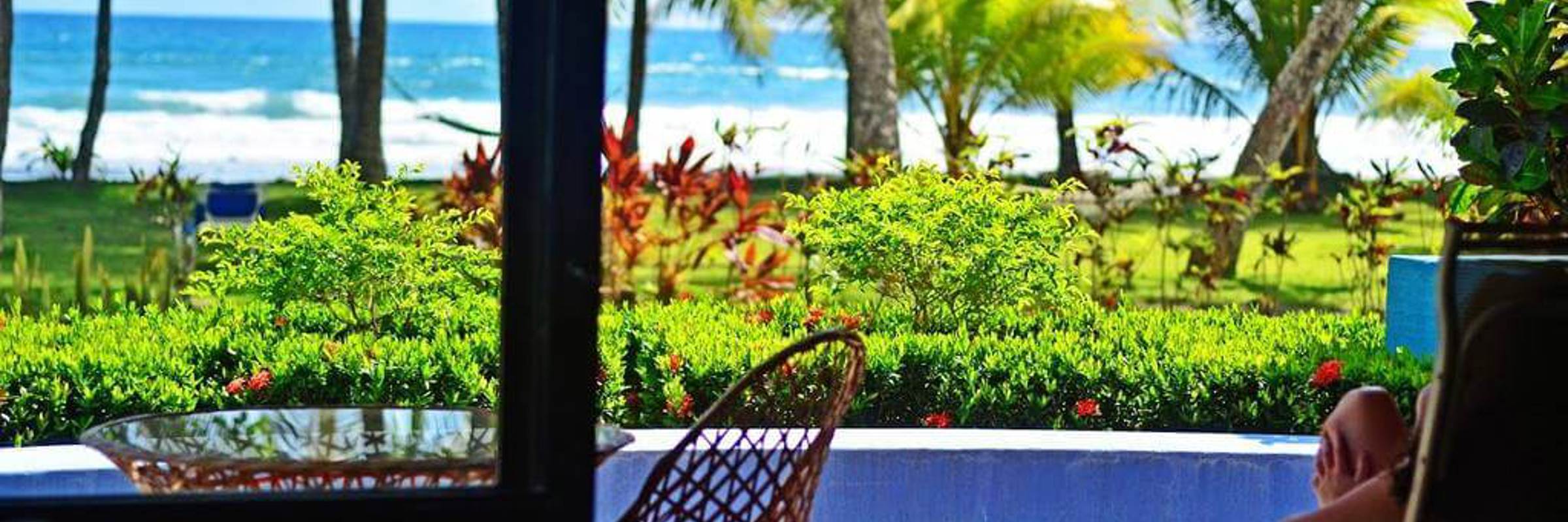 A tourist lounges on a terrace with a view of palm trees, a pool, and Playa Esterillos Beach in Costa Rica. 