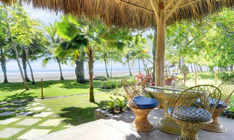 Thatched-roof beach hut with rustic wicker chairs and table, overlooking palm trees and the Pacific Ocean at Playa Esterillos. 