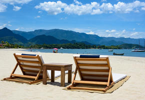 Wooden loungers on the white sand beach of Angra dos Reis, with boats on calm waters and forested mountains beyond. 