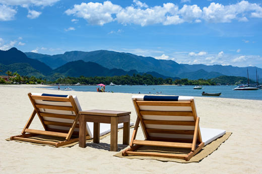 Wooden loungers on the white sand beach of Angra dos Reis, with boats on calm waters and forested mountains beyond. 