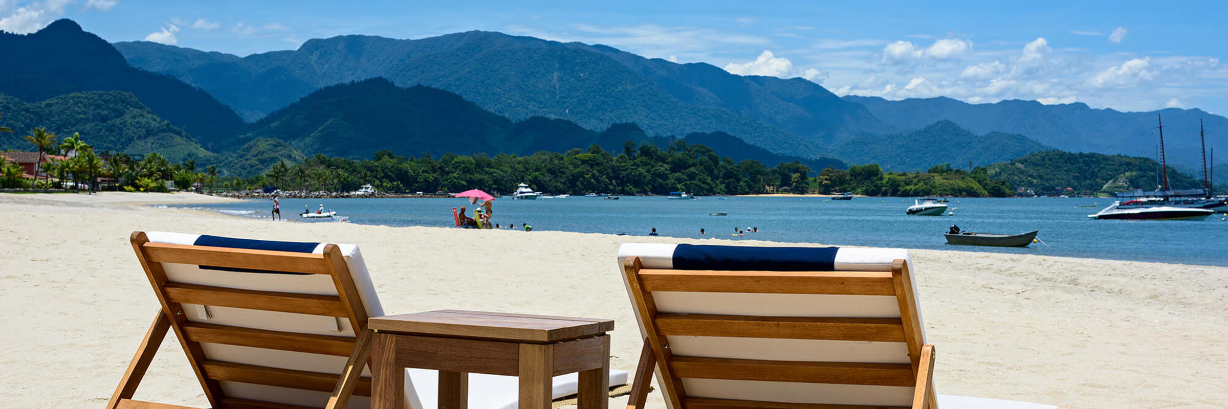 Wooden loungers on the white sand beach of Angra dos Reis, with boats on calm waters and forested mountains beyond. 