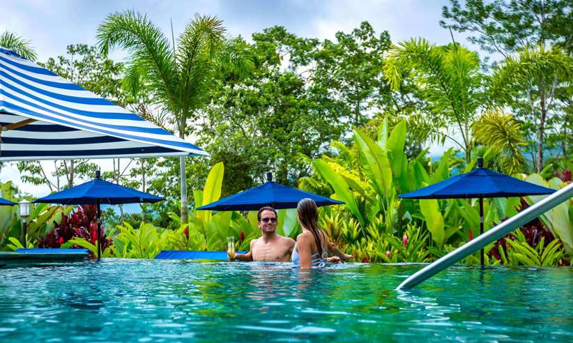 Couple enjoying a tropical escape in the infinity pool at Nayara Springs, surrounded by lush greenery and vibrant umbrellas. 