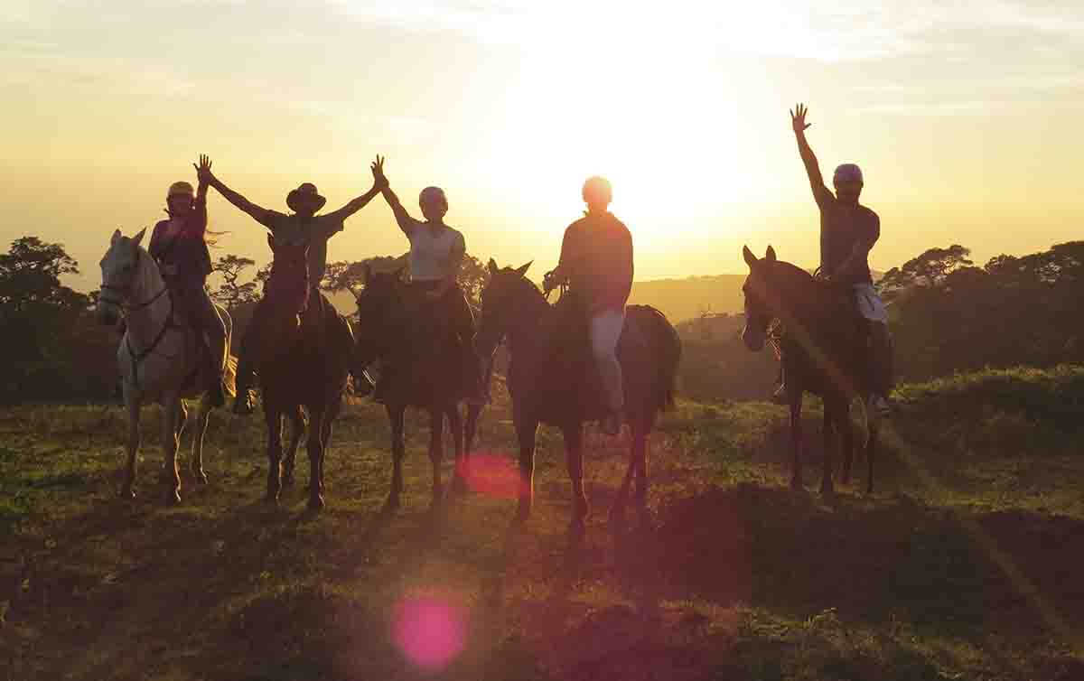 Silhouetted riders on horseback raise their arms against a golden Costa Rican sunset on an open grassy field. 