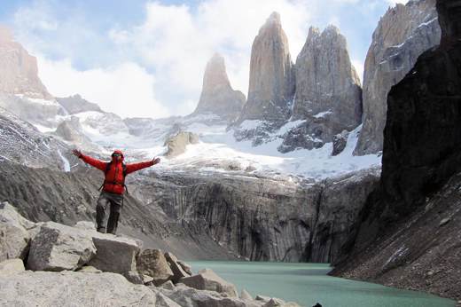 Hiker in red jacket celebrates at the iconic Base of the Torres del Paine with granite spires and turquoise lake in the background. 