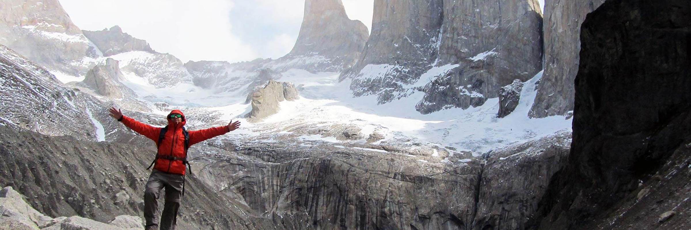 Hiker in red jacket celebrates at the iconic Base of the Torres del Paine with granite spires and turquoise lake in the background. 