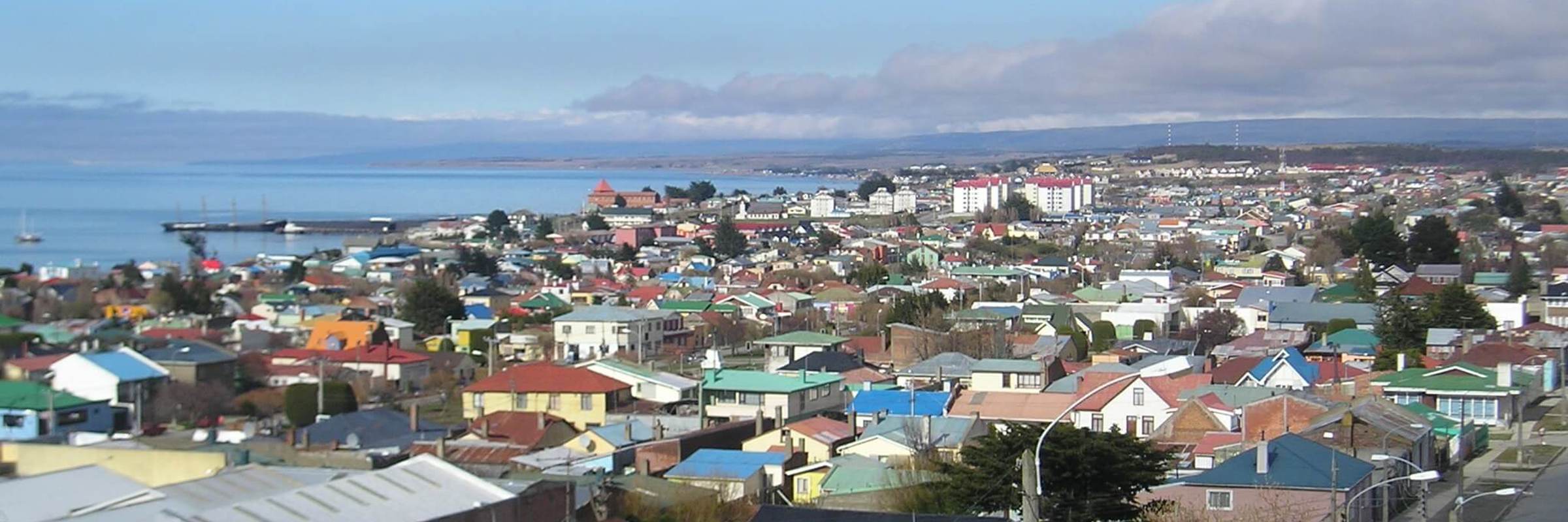 A panoramic view of colourful rooftops in Punta Arenas with the Straight of Magellan stretching out beneath a crisp blue sky. 