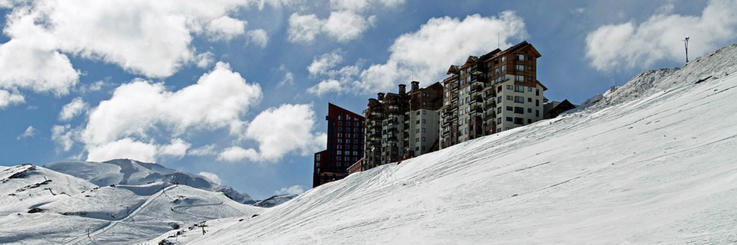 A ski resort building stands on a snowy slope beneath a sky dotted with fluffy clouds in Valle Nevado, Chile. 