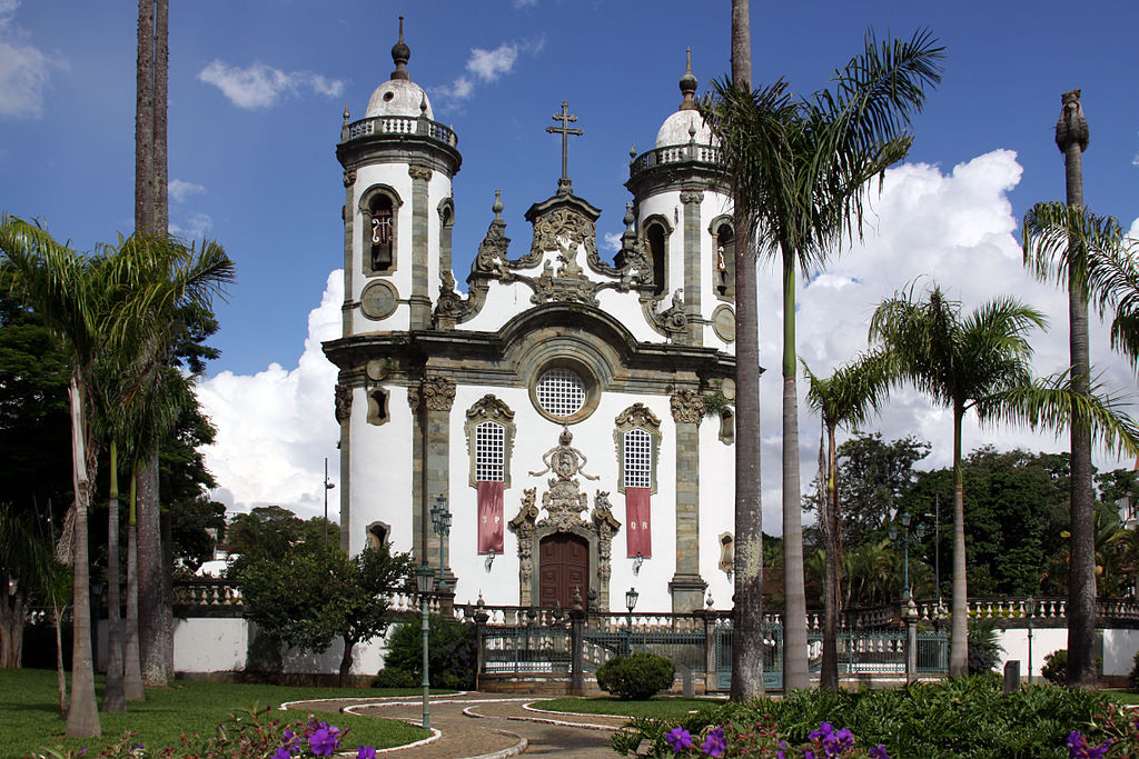 Baroque church in Sao Joao del Rei, Brazil, framed by palm trees and gardens under a bright blue sky. 