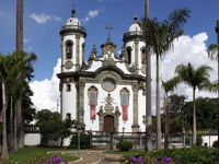 Baroque church in Sao Joao del Rei, Brazil, framed by palm trees and gardens under a bright blue sky. 