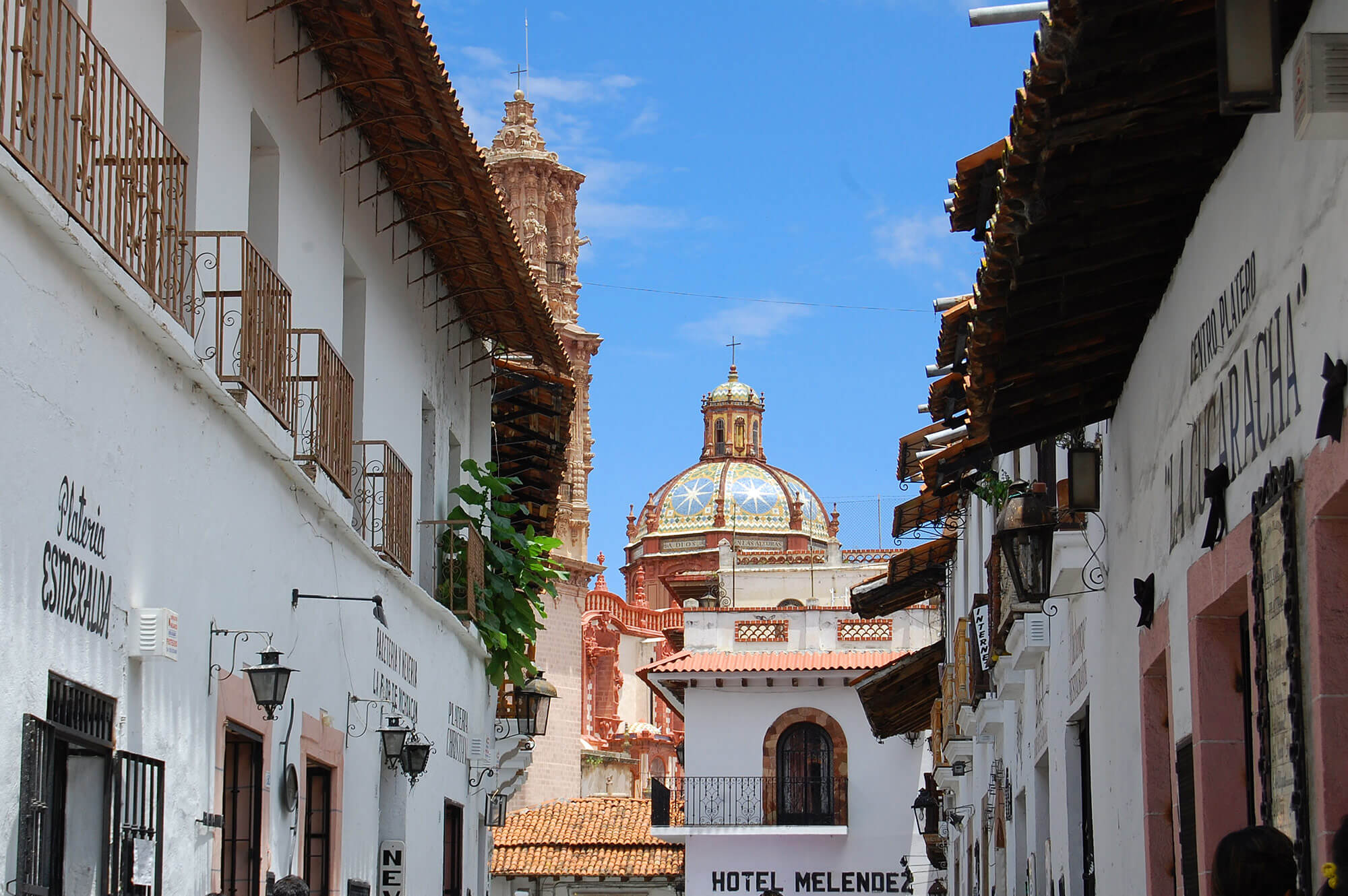 Whitewashed colonial street with balconies, lanterns, and a distant church dome in Taxco, Mexico. 