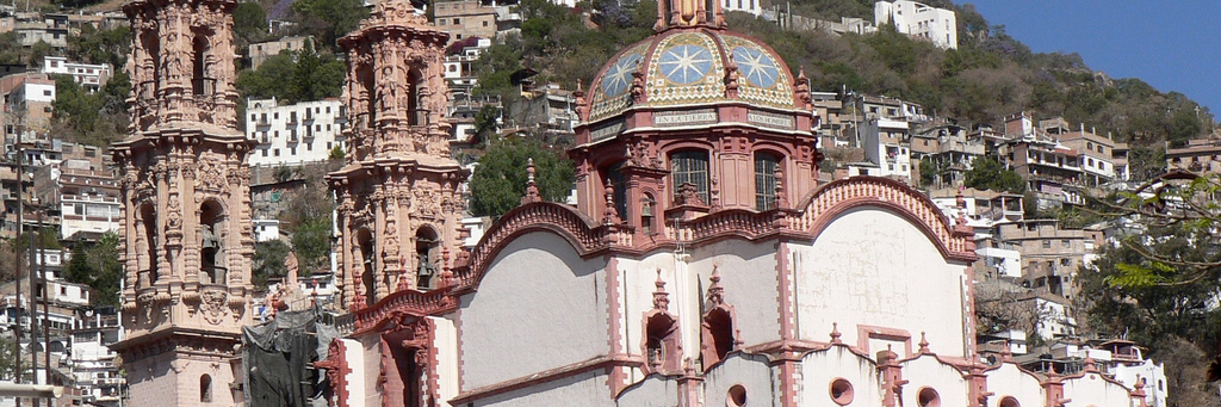 View of Santa Prisca Church in Taxco, a colonial silver town in Mexico, surrounded by hillside homes and scenic views. 