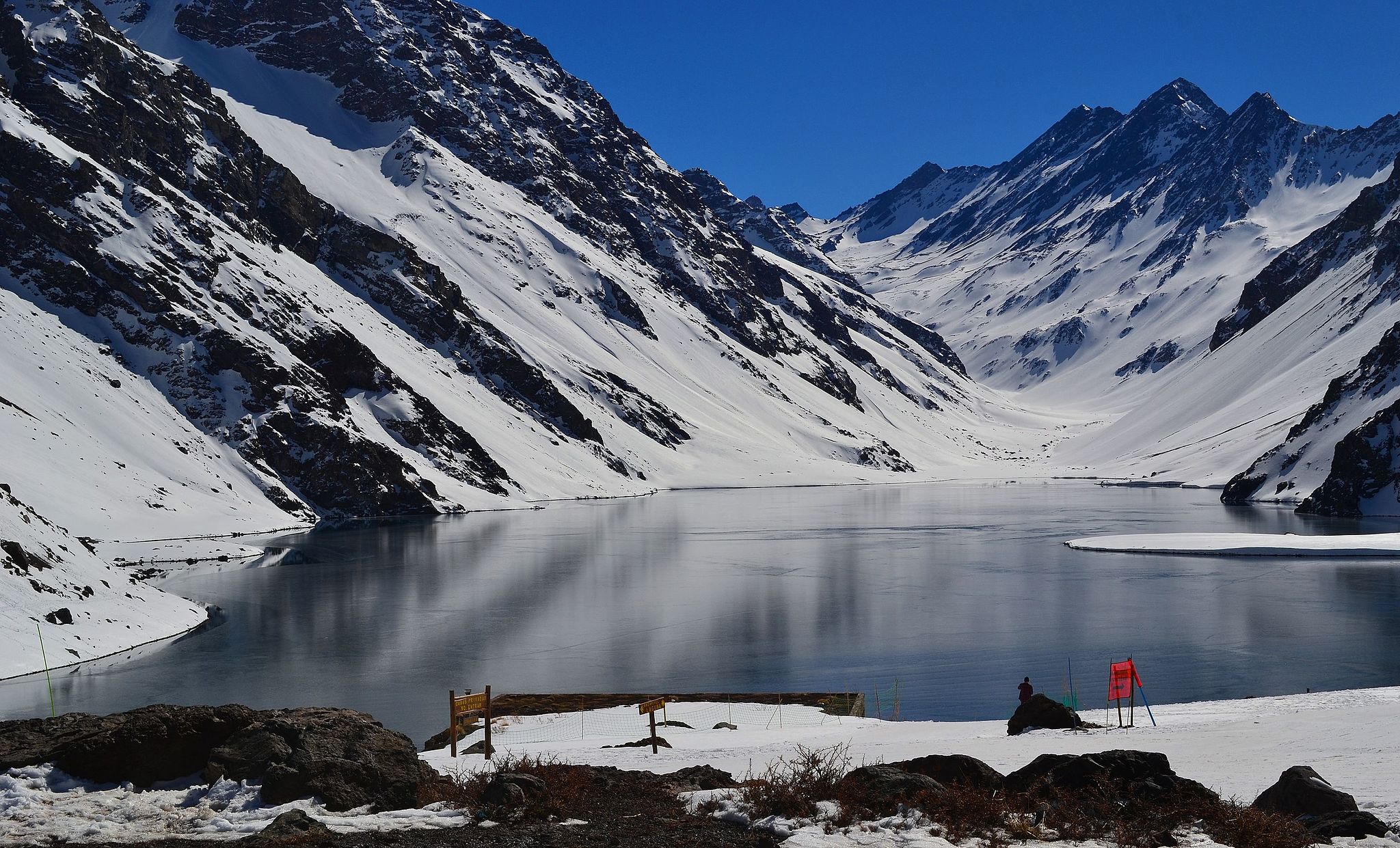 Snow-covered Andes mountains reflect in a still alpine lake near Portillo, Chile, under a clear deep-blue sky. 