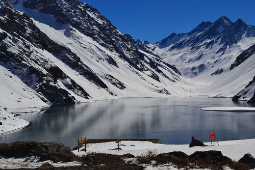 Snow-covered Andes mountains reflect in a still alpine lake near Portillo, Chile, under a clear deep-blue sky. 