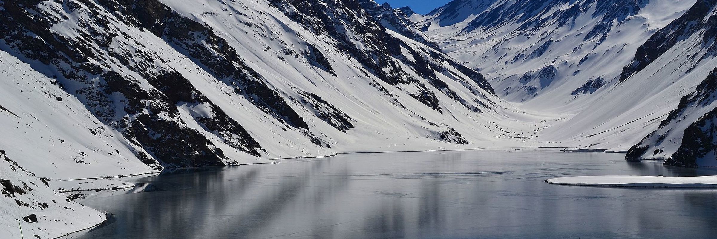 Snow-covered Andes mountains reflect in a still alpine lake near Portillo, Chile, under a clear deep-blue sky. 