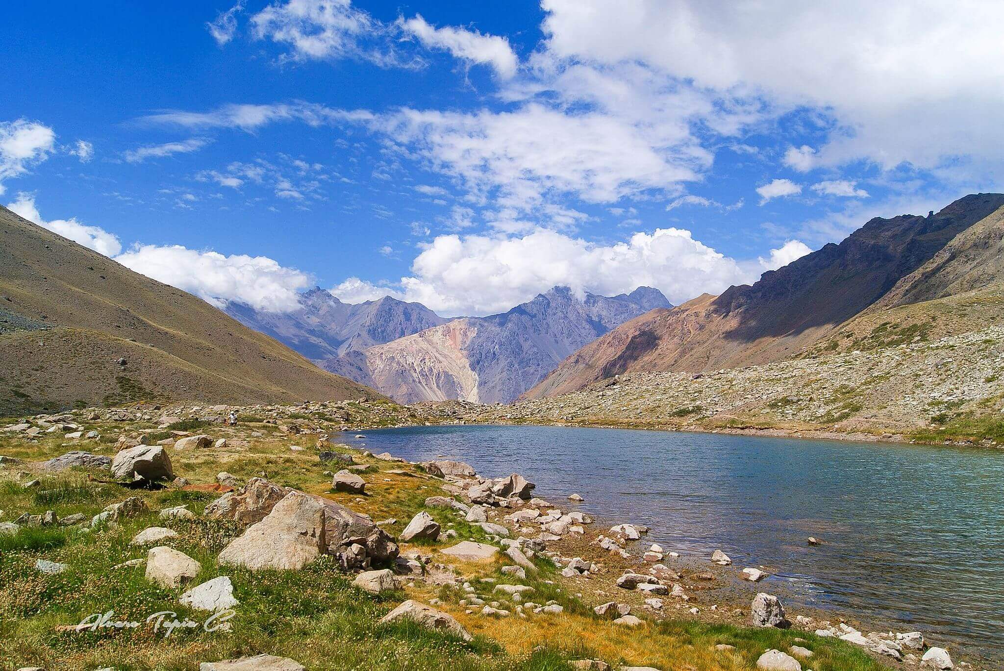 Clear river runs through the rocky Cajon del Maipo valley, framed by rugged peaks and a bright blue sky in the Andes. 