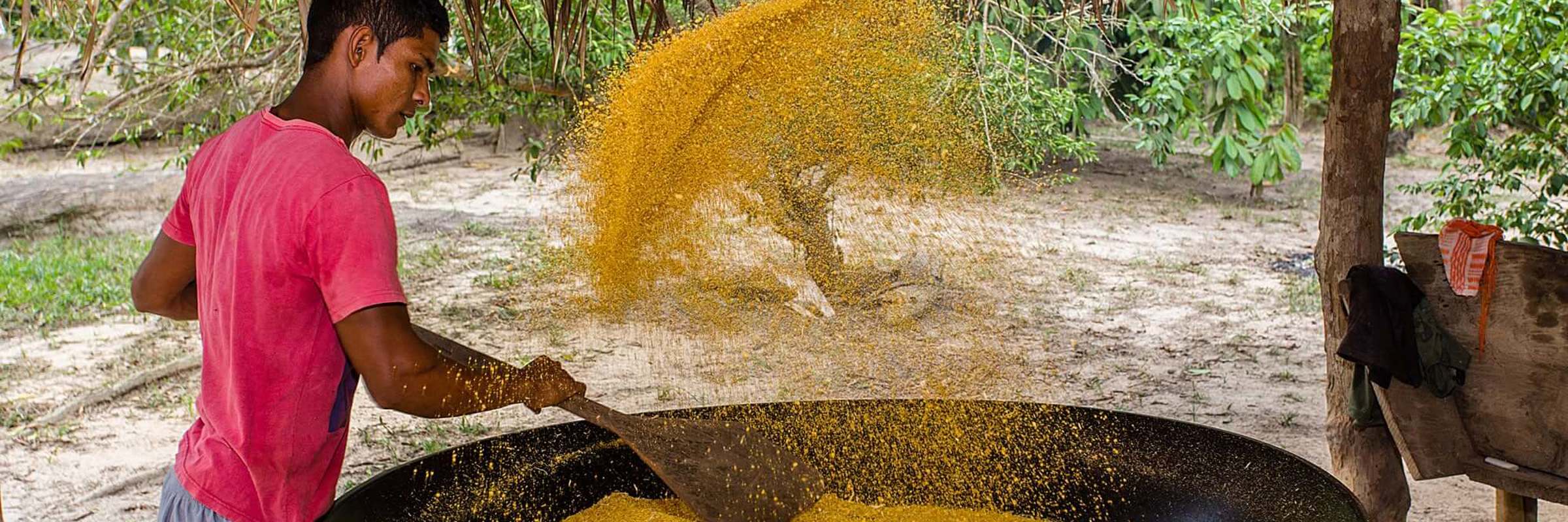Local Amazonian man preparing farofa over a large open pan under a thatched roof in a traditional village setting. 