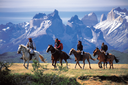Riders on horseback traverse the Patagonian plains with Torres del Paine's jagged peaks rising behind. 