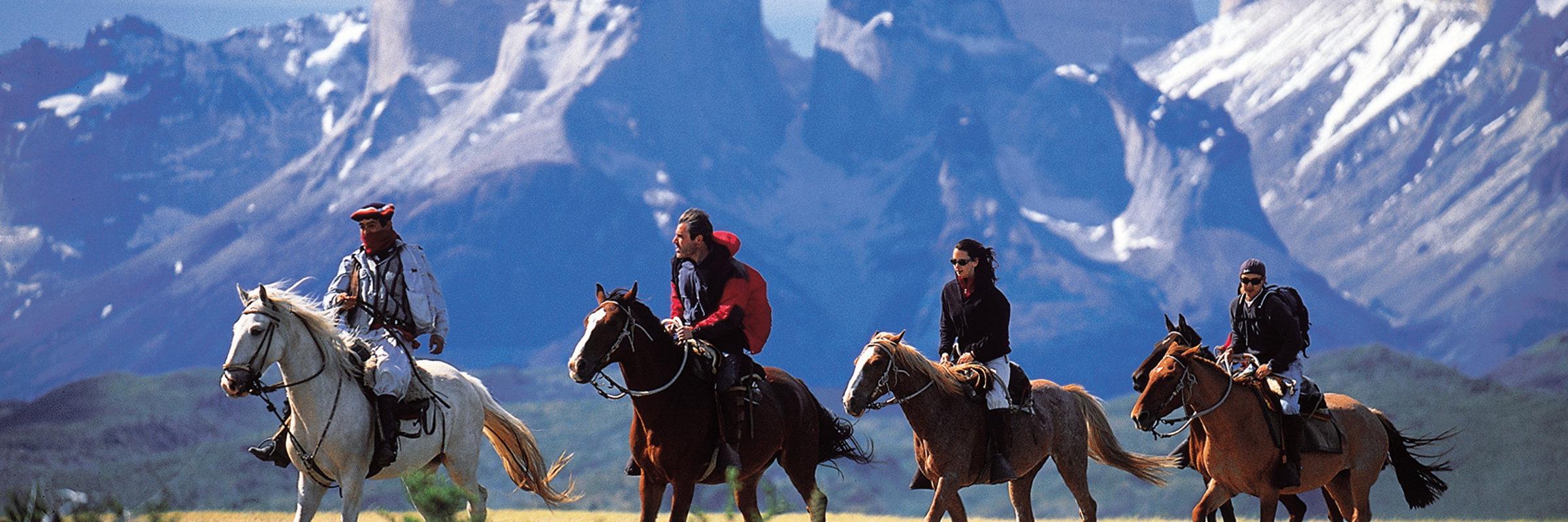 Riders on horseback traverse the Patagonian plains with Torres del Paine's jagged peaks rising behind. 