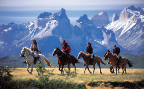 Riders on horseback traverse the Patagonian plains with Torres del Paine's jagged peaks rising behind. 
