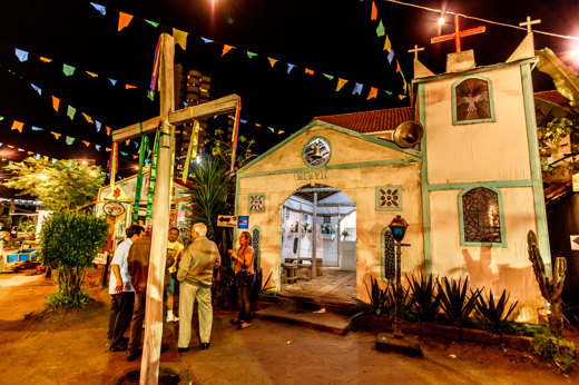 Colourful June Festival night scene with a chapel, wooden cross, and hanging flags in Brazil's Festa Junina. 