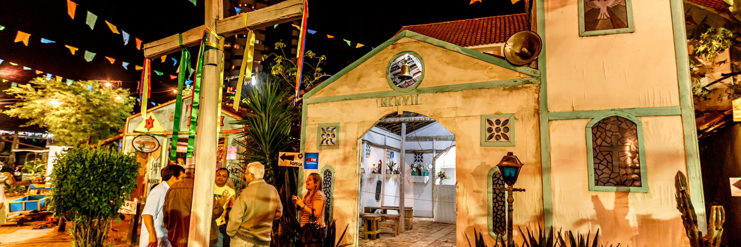 Colourful June Festival night scene with a chapel, wooden cross, and hanging flags in Brazil's Festa Junina. 