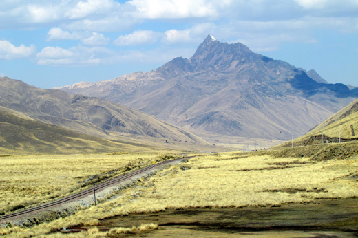 A quiet road winds through the rugged Peruvian Andes under a brilliant blue sky. 