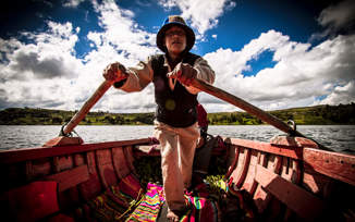 A traditional boatman rowing on Lake Titicaca, Peru, wearing local attire on a wooden boat under a bright sky. 
