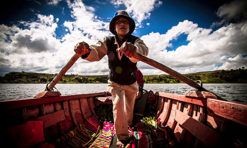 A traditional boatman rowing on Lake Titicaca, Peru, wearing local attire on a wooden boat under a bright sky. 