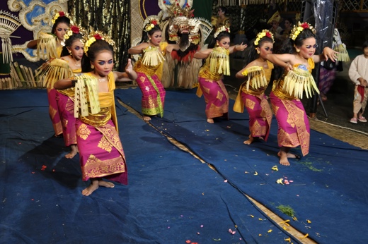 Young Balinese girls in vibrant costumes perform a traditional dance with elegant gestures on a decorated stage. 